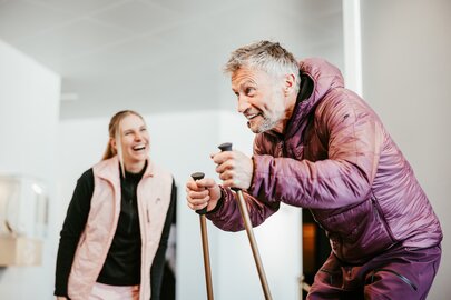 Ein Mann im Skianzug trainiert drinnen mit Skistöcken, während eine Frau lacht.