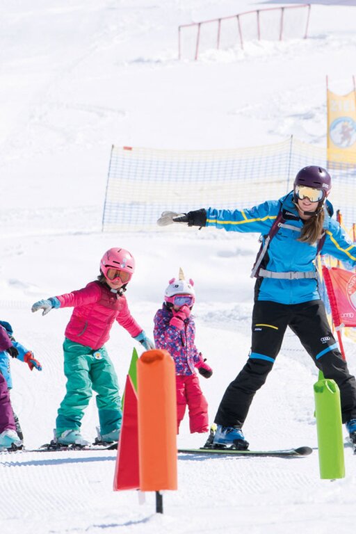 Eine Skilehrerin zeigt Kindern Übungen auf einer Piste.