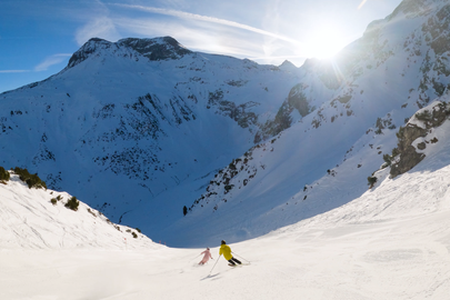 Zwei Personen beim Skifahren auf einer sonnigen Bergpiste mit Blick auf schneebedeckte Gipfel.