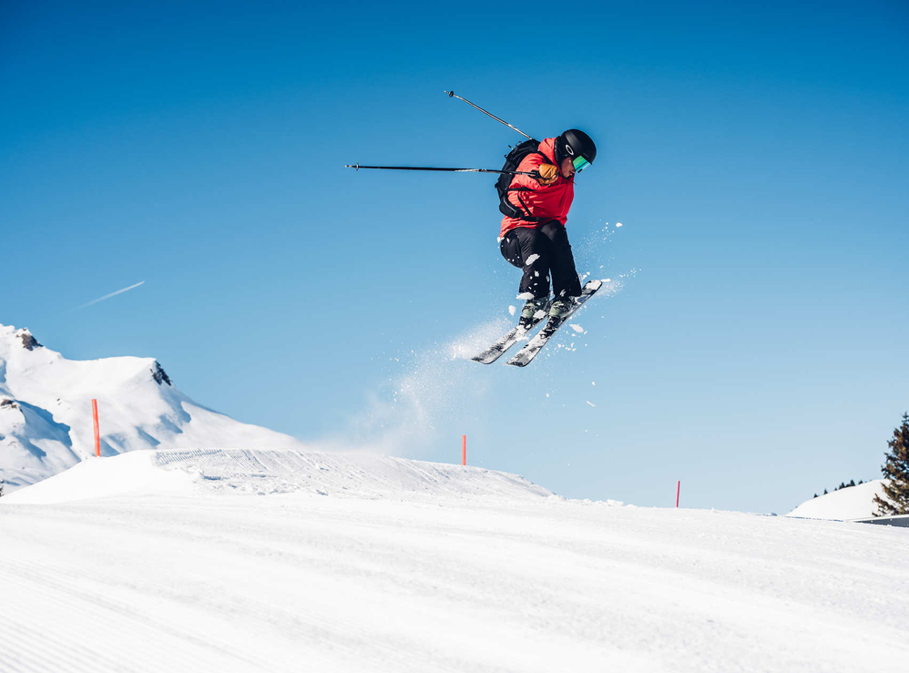 Skifahrer in roter Jacke springt auf verschneiter Piste in die Luft.