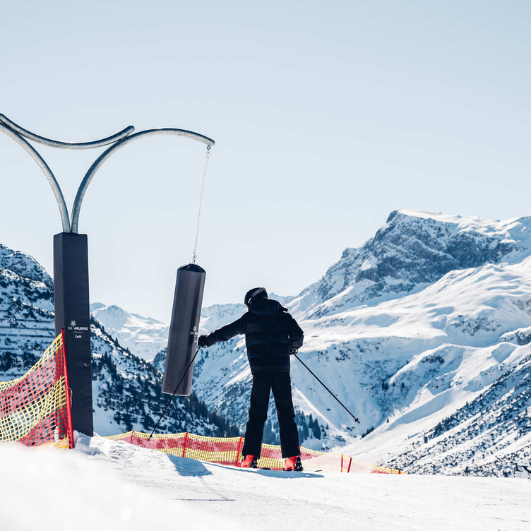 Person auf Ski an der Liftstation mit schneebedeckten Bergen im Hintergrund.