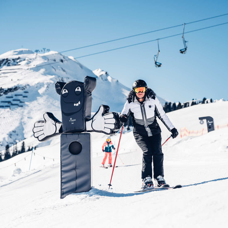 Skifahrer neben einem lustigen, schwarz-weißen Hindernis auf der Piste.