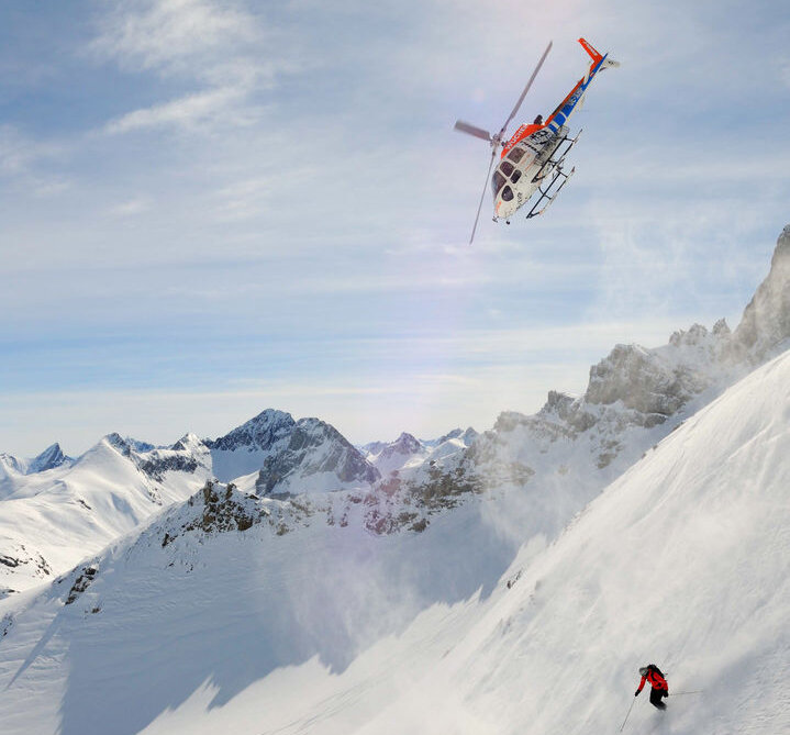 Gruppe von Skifahrern auf steilem Hang, begleitet von einem Hubschrauber im Hintergrund unter sonnigem Himmel.