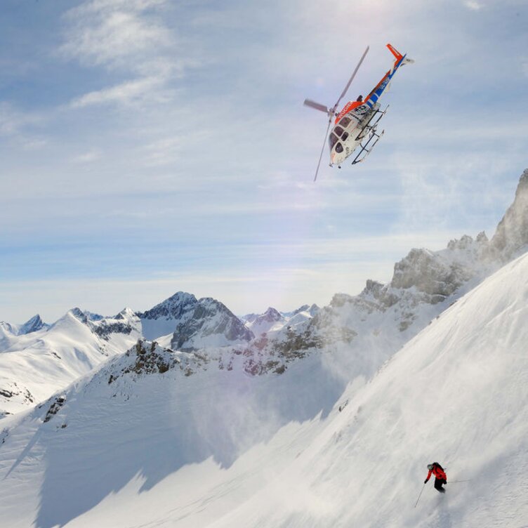 Gruppe von Skifahrern auf steilem Hang, begleitet von einem Hubschrauber im Hintergrund unter sonnigem Himmel.
