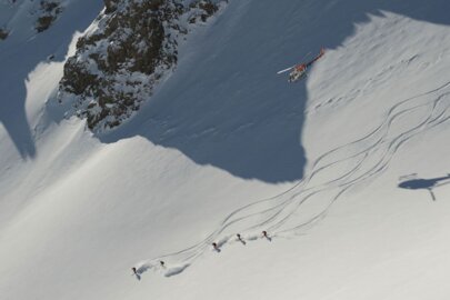 Mehrere Skifahrer fahren einen Steilhang hinunter und werden von einem Hubschrauber begleitet.