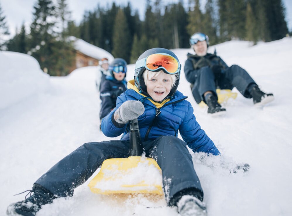 Kinder und Erwachsene rodeln lachend im Schnee.