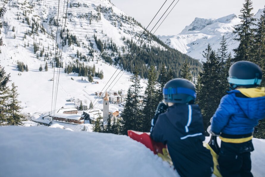 Zwei Kinder mit Helmen sitzen im Schnee mit Blick auf ein verschneites Dorf.