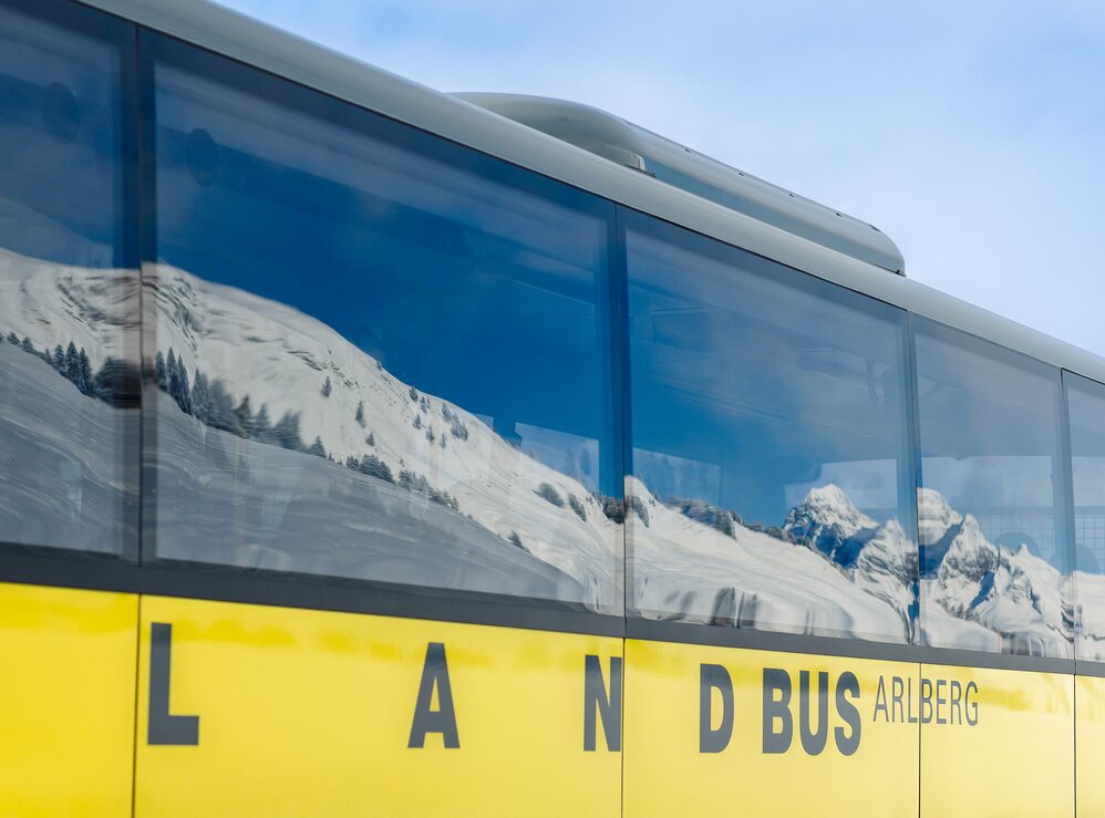 Ein gelber Bus mit der Aufschrift 'Landbus Arlberg' und im Fenster spiegeln sich schneebedeckte Alpen.