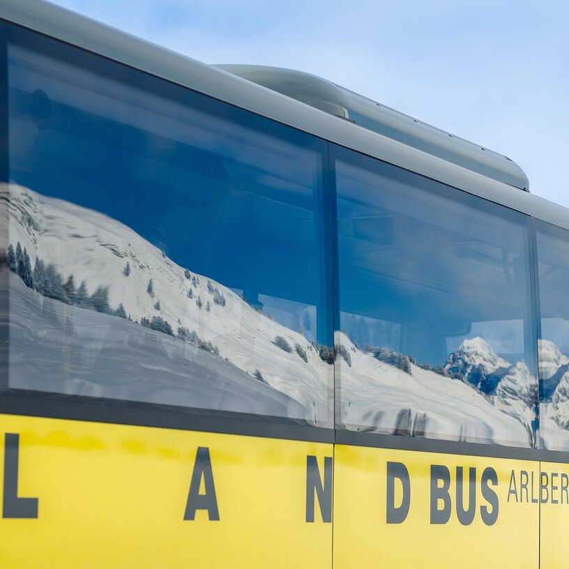 Ein gelber Bus mit der Aufschrift 'Landbus Arlberg' und im Fenster spiegeln sich schneebedeckte Alpen.
