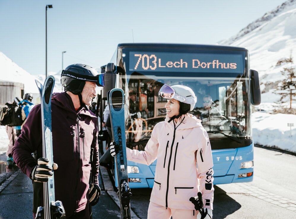 Zwei Personen stehen samt Skiausrüstung vor dem Lech-Dorfbus 703.