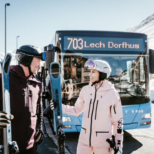 Zwei Personen stehen samt Skiausrüstung vor dem Lech-Dorfbus 703.