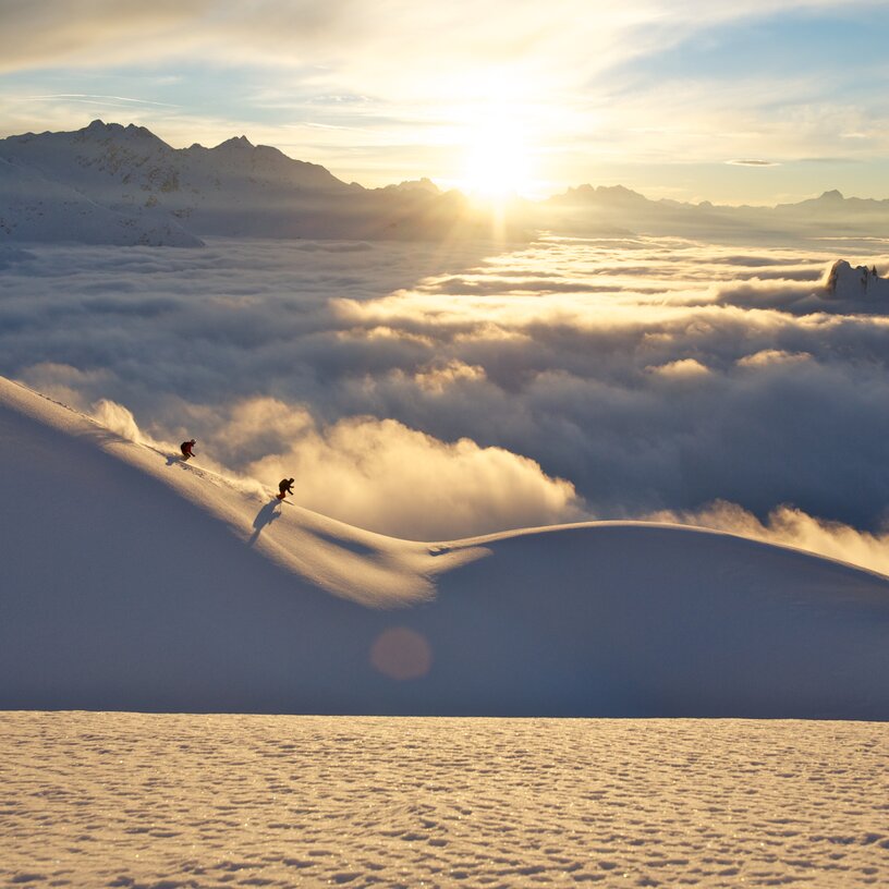 Zwei Skifahrer fahren bei Sonnenaufgang einen verschneiten Hang hinunter, umgeben von Wolken und Bergspitzen.