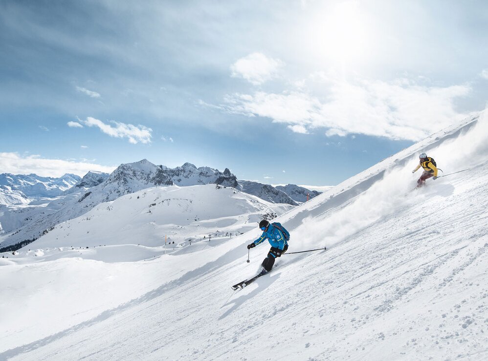 Zwei Skifahrer sausen bei Sonnenschein die Piste hinunter.