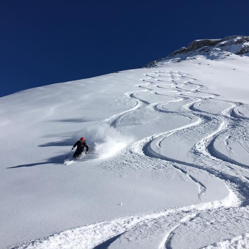 Ein Skifahrer zieht elegante Schwünge in frischem Pulverschnee auf einer steilen Abfahrt unter klarem, blauem Himmel.