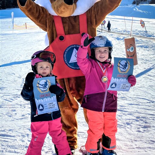 Zwei Kinder in Skiausrüstung freuen sich zusammen mit dem Ski-Maskottchen Foxy über ihre Urkunden.