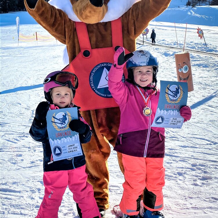Zwei Kinder in Skiausrüstung freuen sich zusammen mit dem Ski-Maskottchen Foxy über ihre Urkunden.
