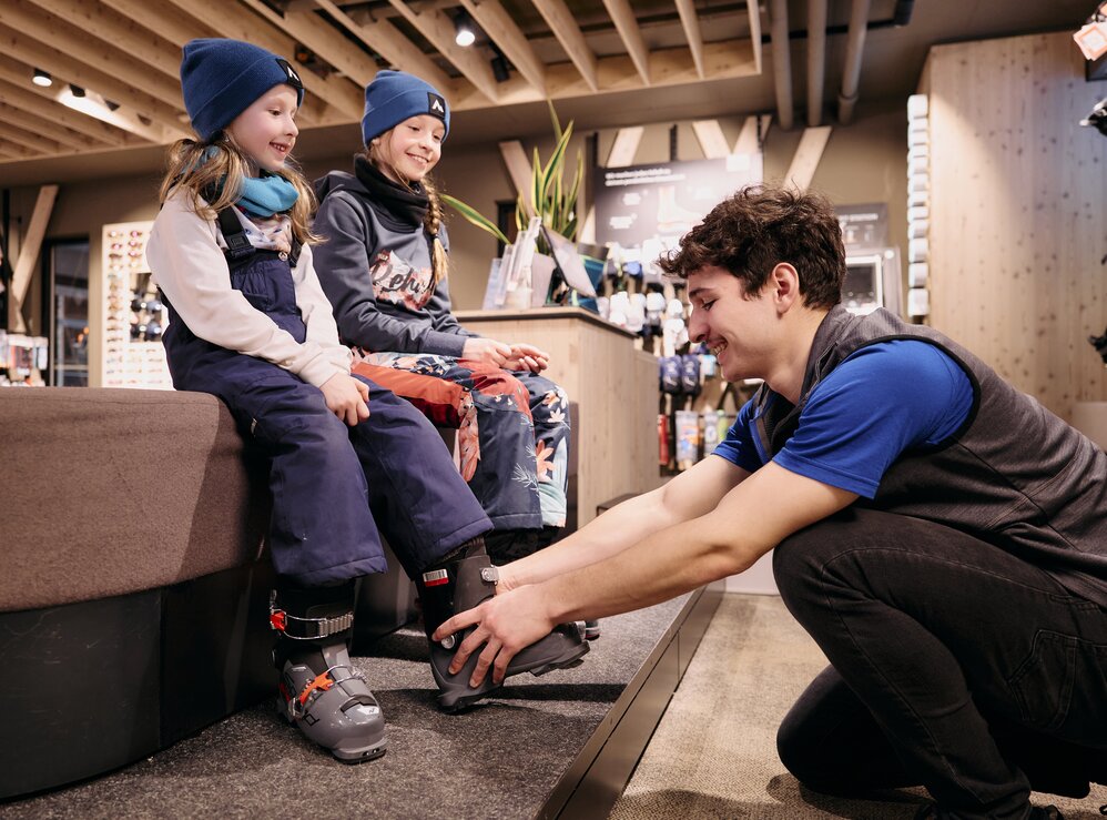 Zwei Kinder in Schneekleidung probieren Skischuhe in einem Geschäft an.