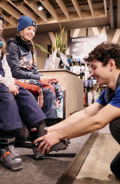 Zwei Kinder in Schneekleidung probieren Skischuhe in einem Geschäft an.