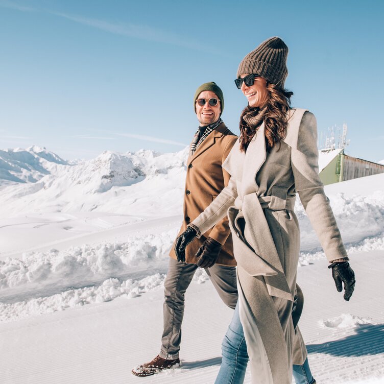 Ein Paar spaziert stilvoll gekleidet in einer verschneiten Berglandschaft.