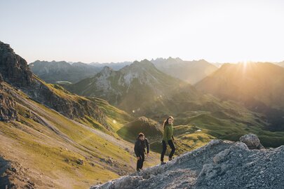 Zwei Personen wandern bei Sonnenaufgang durch bergiges Gelände.