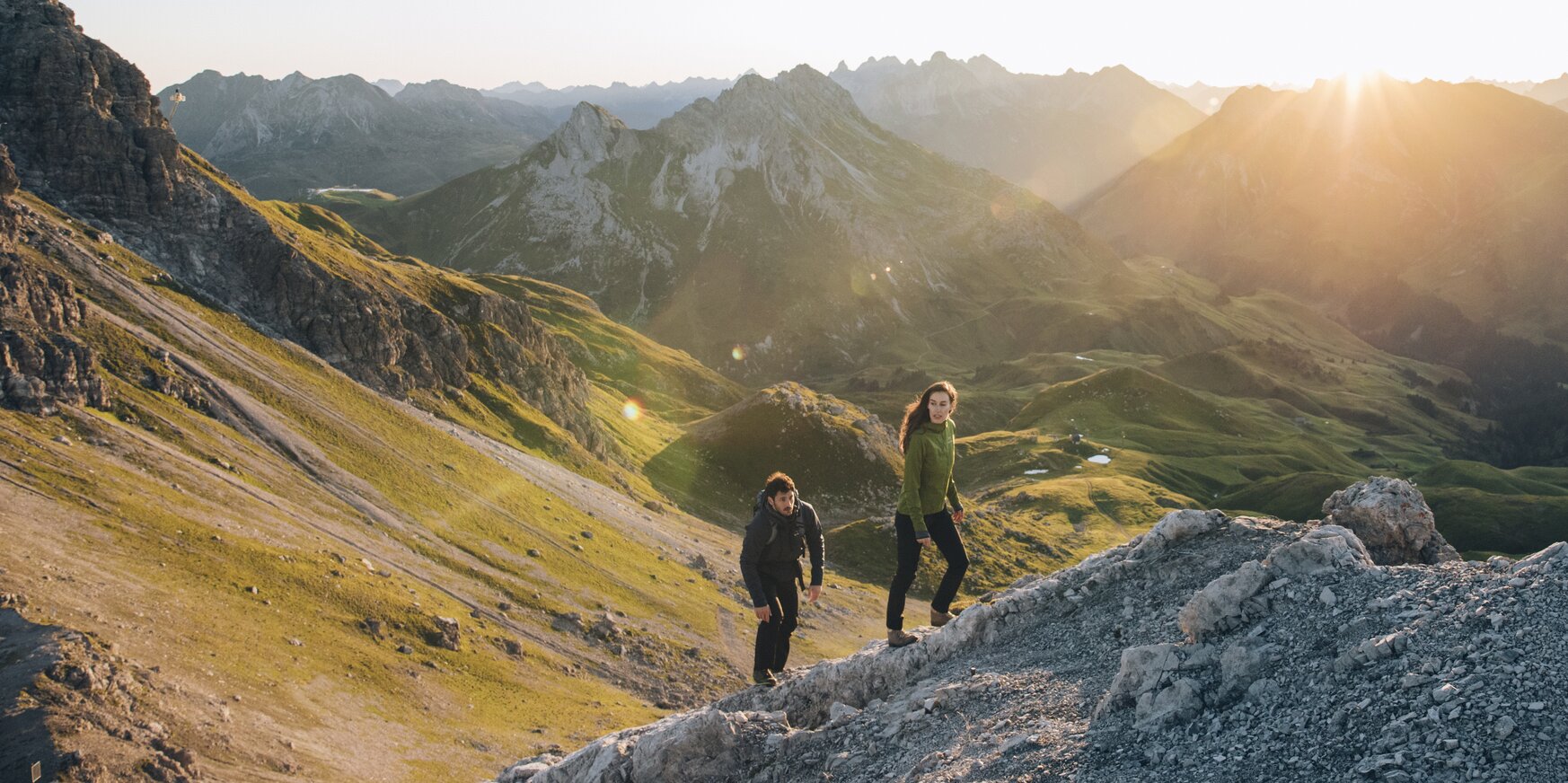 Zwei Personen wandern bei Sonnenaufgang durch bergiges Gelände.