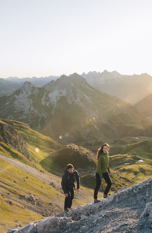 Zwei Personen wandern bei Sonnenaufgang durch bergiges Gelände.