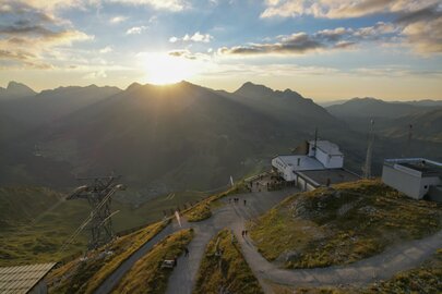 Menschen genießen den Sonnenuntergang auf einem Berg mit Seilbahn und Hütten im Hintergrund.