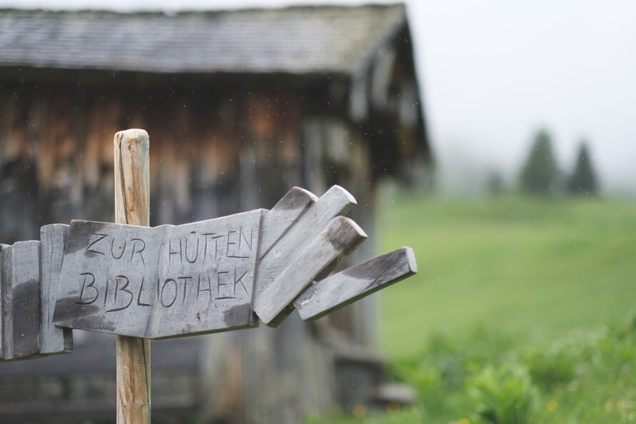 Holzwegweiser zur Hüttenbibliothek, mit grüner Wiese im Hintergrund.