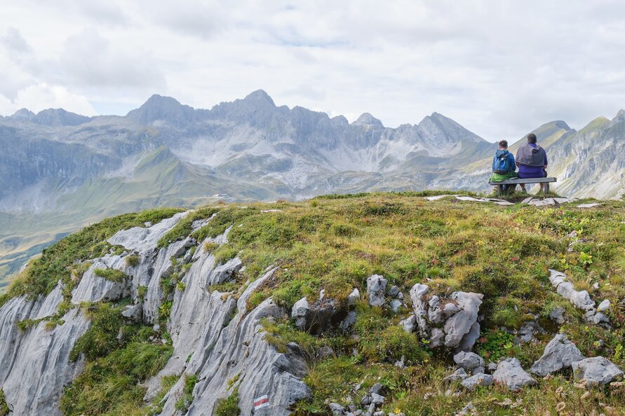 Zwei Personen sitzen auf einer Bank und blicken auf das umliegende Bergpanorama.