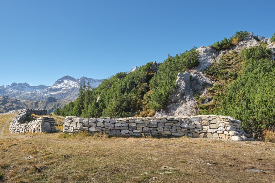 Steinmauer in alpiner Landschaft mit Bergen und Vegetation.