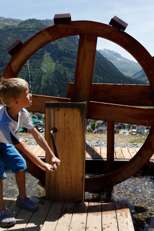 Junge spielt auf dem Wasserspielplatz Kriegerhorn mit einem Wasserrad mit Bergpanorama im Hintergrund.