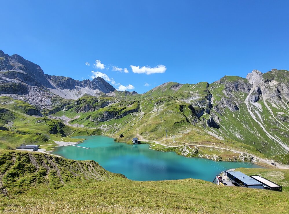 Blick auf den türkisfarbenen Zürsersee umgeben von den grünen Bergen am Naturberg Seekopf unter blauem Himmel.