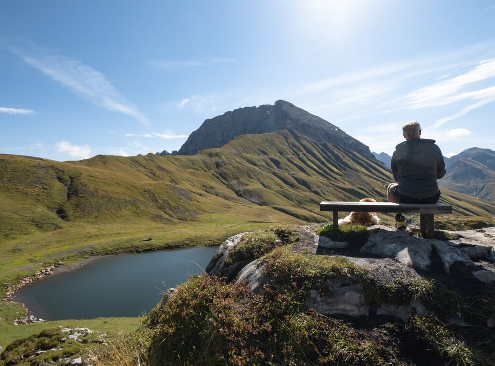 Ein Wanderer sitzt auf einer Bank und genießt die Aussicht auf den Bergsee am Panoramaberg Rüfikopf.