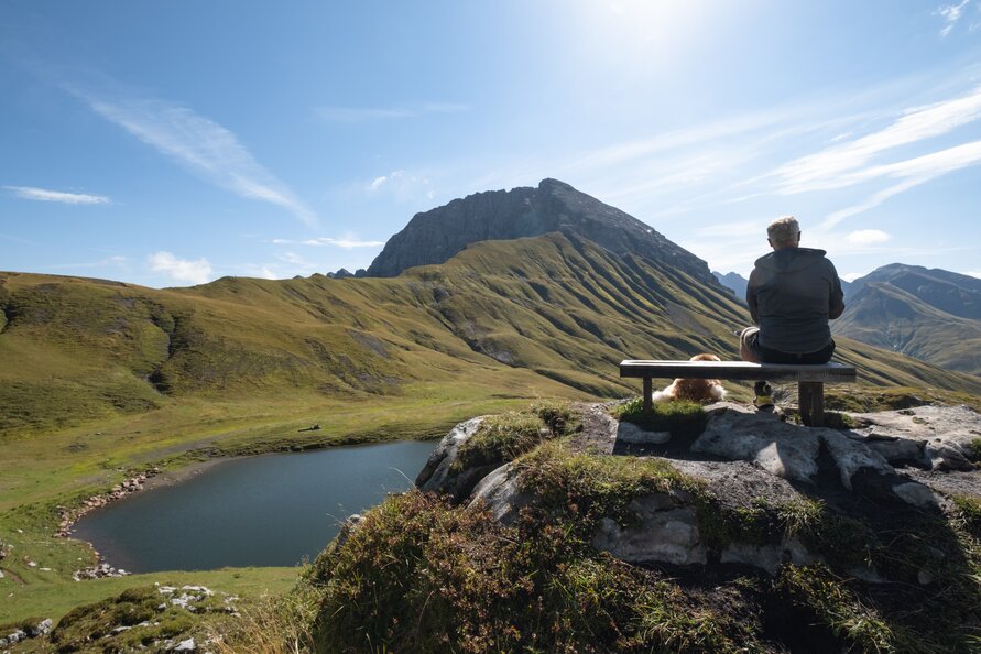 Ein Wanderer sitzt auf einer Bank und genießt die Aussicht auf den Bergsee am Panoramaberg Rüfikopf.