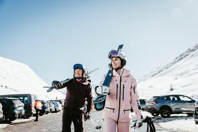 Two people in ski attire carrying skis through snowy landscape.