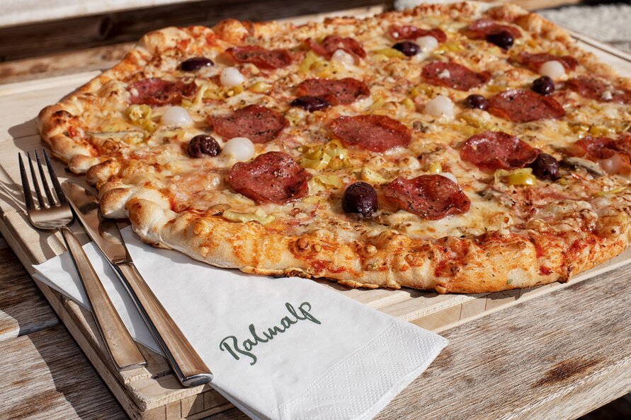 Große Hüttenpizza mit Salami, Oliven und Zwiebeln auf Holzbrett, mit Besteck und Balmalp-Serviette daneben.