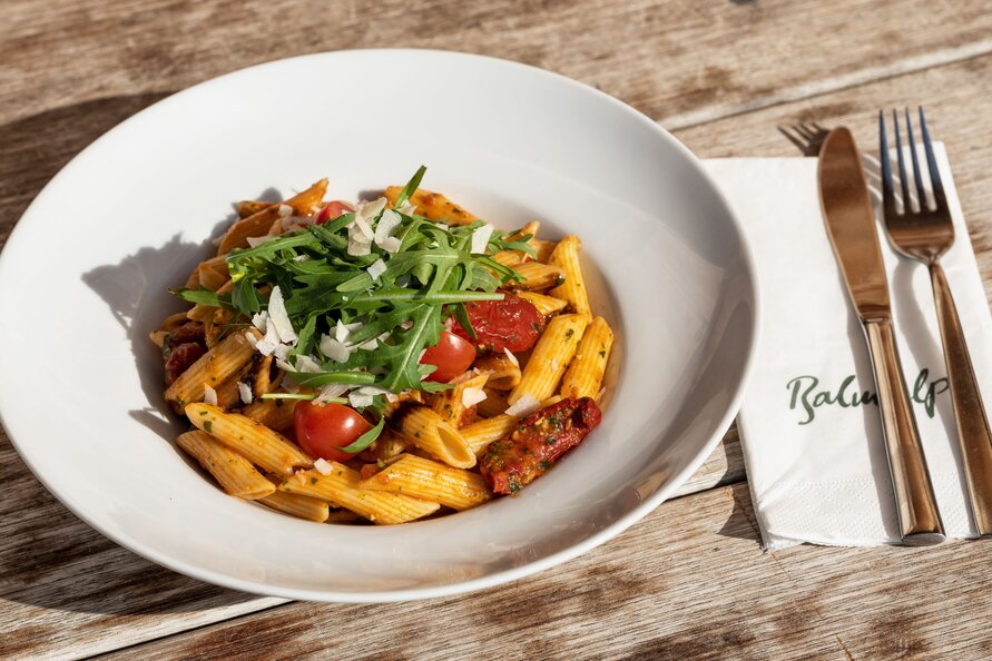 Ein Teller Penne Arrabbiata mit Rucola und Parmesan auf einem Holztisch, daneben Serviette mit Balmalp-Logo.