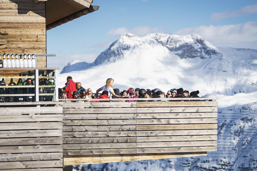 Gruppe von Menschen genießt die Sonne auf der Terrasse der Balmalp, mit schneebedeckten Bergen im Hintergrund.