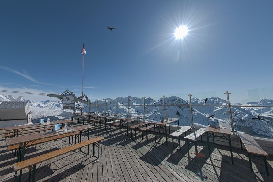 Leere sonnenbeschienene Terrasse des Rüfikopf Restaurants mit schneebedeckten Bergen im Hintergrund.