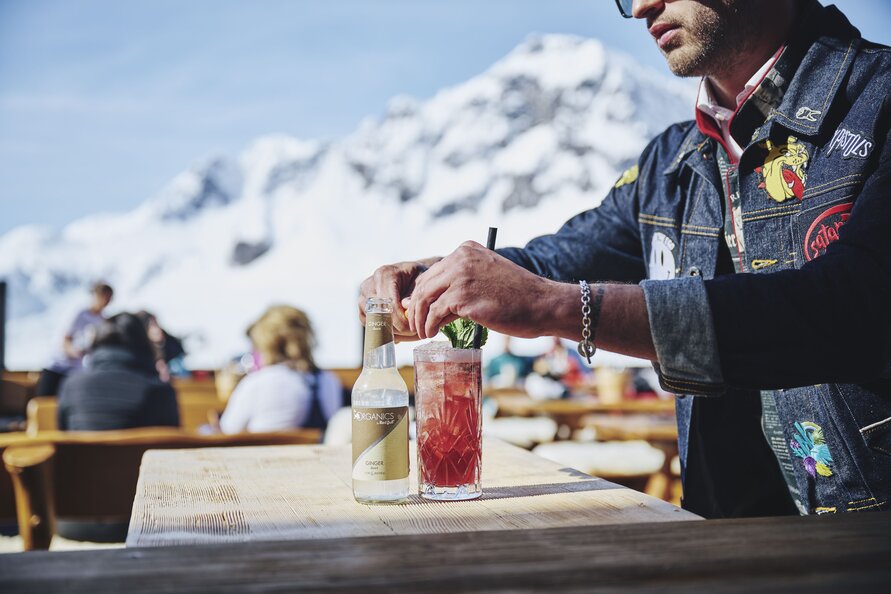 Person bereitet einen Cocktail auf der Terrasse des Restaurants Trittalpe mit Bergblick zu.