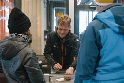 Menschen kaufen Tickets an einem Skischalter in einer Hütte.