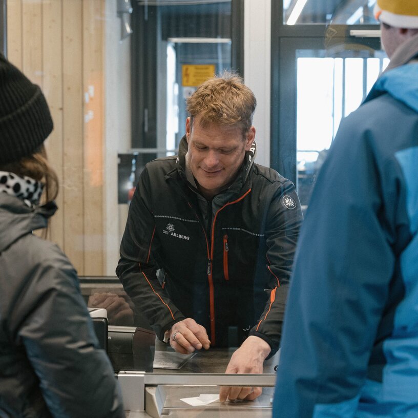 Menschen kaufen Tickets an einem Skischalter in einer Hütte.