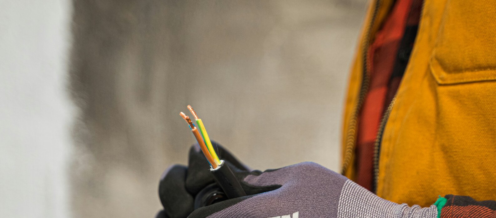 Person mit einer gelben Jacke hält Kabeln in den Händen und trägt dabei Schutzhandschuhe.