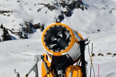 Schneekanone auf verschneitem Berg mit blauem Himmel im Hintergrund.