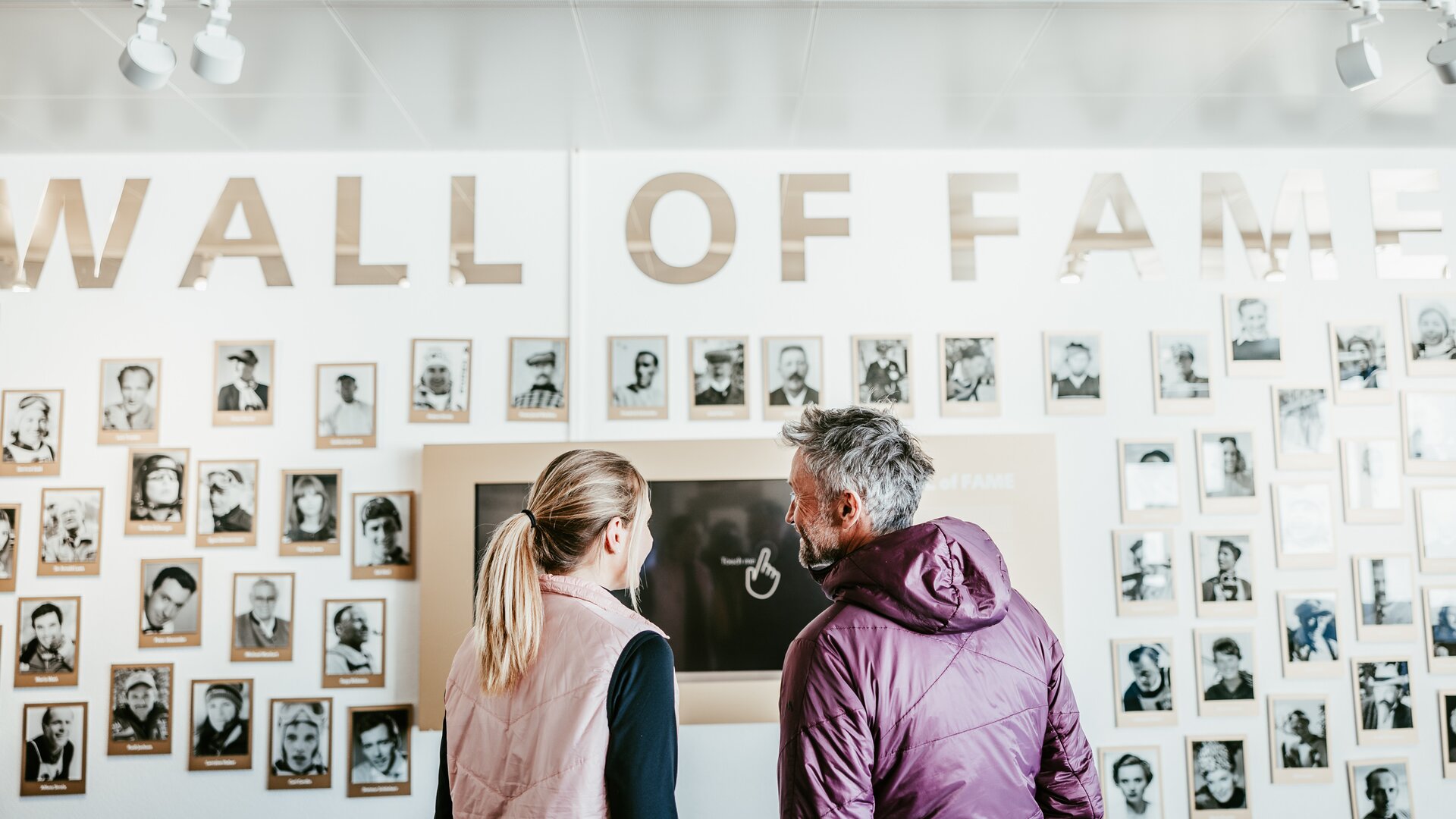 Zwei Personen betrachten eine 'Wall of Fame' mit vielen Fotos von Skifahrer-Legenden.