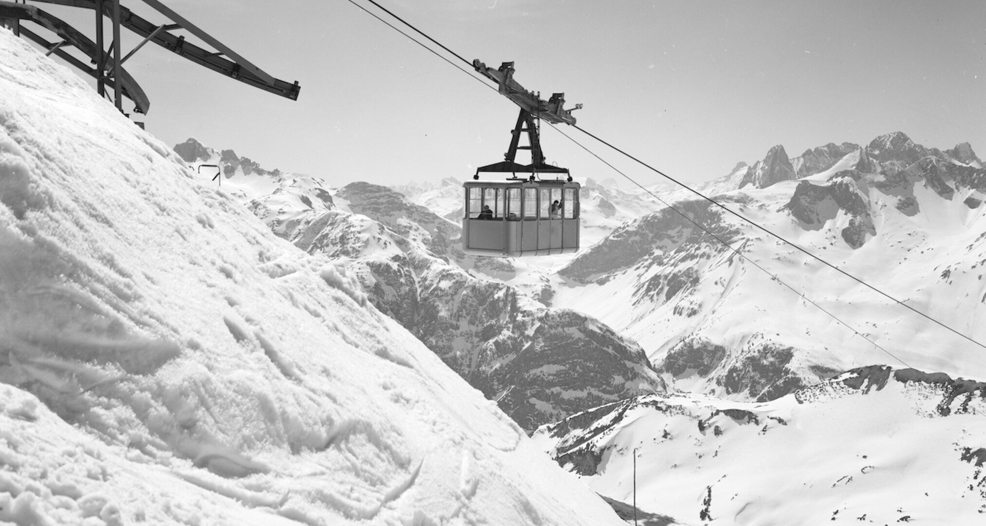 Schwarz-Weiß-Bild einer Seilbahn in den Bergen mit verschneiten Gipfeln im Hintergrund.