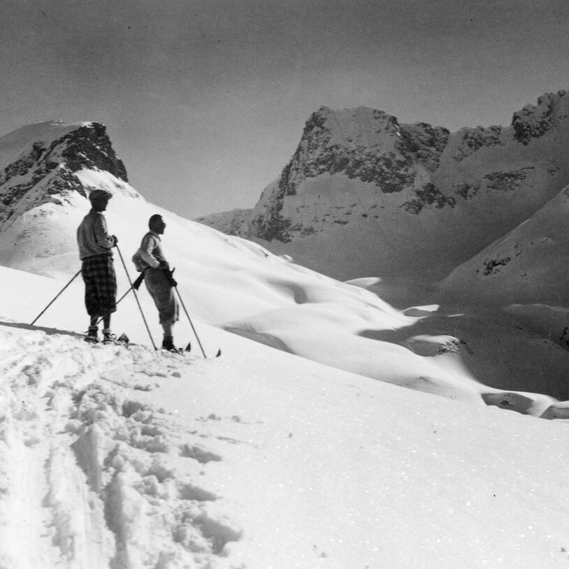 Schwarz-Weiß-Bild: Zwei Skifahrer stehen auf einem verschneiten Hang mit Bergkulisse.