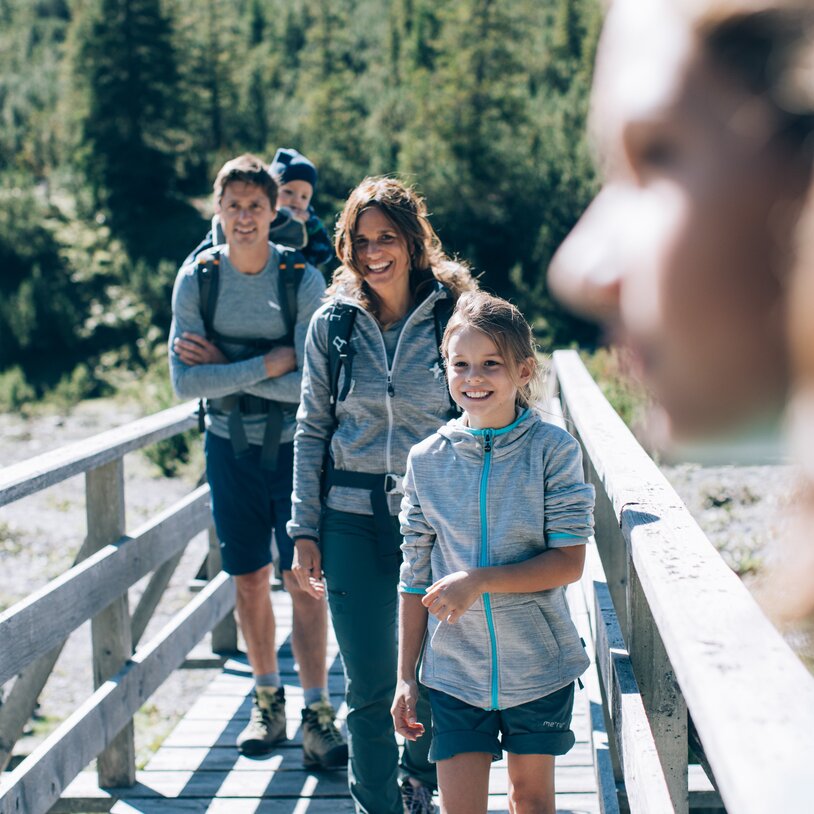 Familie wandert auf Holzbrücke über Fluss mit Wald im Hintergrund.