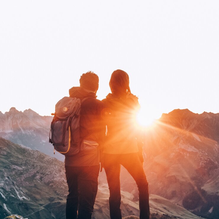 Zwei Personen stehen auf einem Berg und schauen in den Sonnenaufgang.