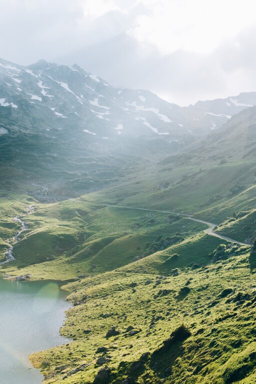Ein Bergsee mit grünen Hügeln im Morgenlicht, Pfad entlang des Ufers.